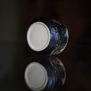 Puede incluir: Un peque&ntilde;o recipiente de cer&aacute;mica redondo con un exterior negro texturizado y una base circular de color beige claro suave que contrasta. El objeto presenta un acabado de superficie rugoso y moteado, destacando su fondo plano sin esmaltar.