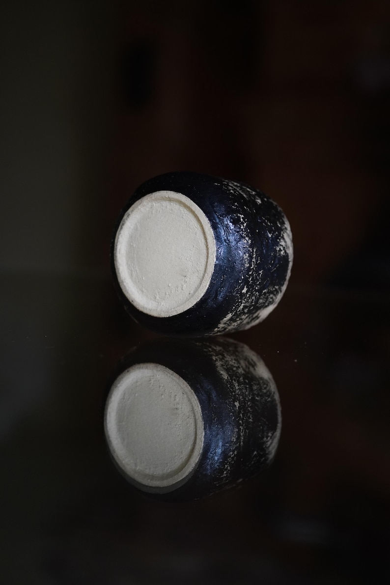 Puede incluir: Un peque&ntilde;o recipiente de cer&aacute;mica redondo con un exterior negro texturizado y una base circular de color beige claro suave que contrasta. El objeto presenta un acabado de superficie rugoso y moteado, destacando su fondo plano sin esmaltar.