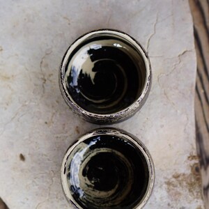 Puede incluir: Dos tazas de cer&aacute;mica peque&ntilde;as y redondas con un acabado exterior oscuro y moteado. El interior de cada taza presenta un patr&oacute;n de remolinos pintado a mano con esmalte negro y crema, creando un efecto de movimiento circular distintivo.