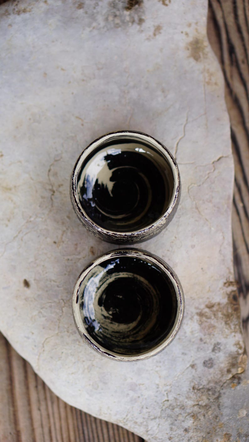 Puede incluir: Dos tazas de cer&aacute;mica peque&ntilde;as y redondas con un acabado exterior oscuro y moteado. El interior de cada taza presenta un patr&oacute;n de remolinos pintado a mano con esmalte negro y crema, creando un efecto de movimiento circular distintivo.