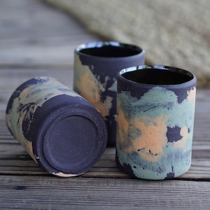 May include: Three handmade ceramic tumblers with a dark blue base and a speckled design in shades of orange and green. The tumblers are cylindrical with a smooth, matte finish and a dark interior. They are arranged on a wooden surface.