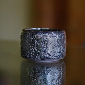 May include: A small, dark grey ceramic bowl with a textured, rough surface. The bowl is cylindrical with a slightly rounded rim and sits on a reflective surface. The bowl's exterior has a unique, organic appearance.