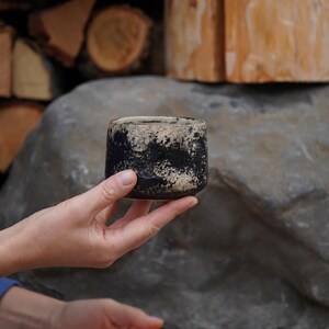 Puede incluir: Una peque&ntilde;a taza de cer&aacute;mica cil&iacute;ndrica con un acabado texturizado y moteado en un patr&oacute;n de alto contraste negro y blanco roto. El recipiente tiene un borde grueso e irregular y una base redondeada, mostrando un aspecto artesanal.