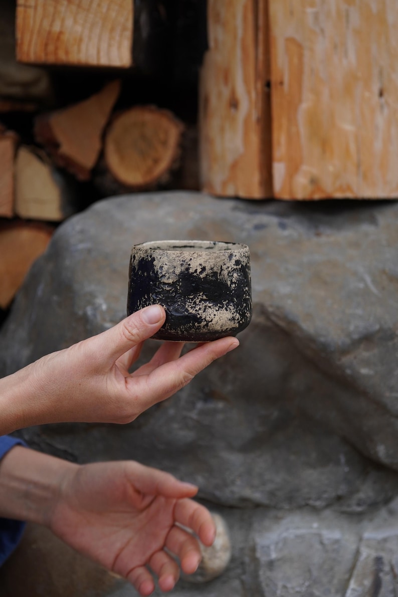 Puede incluir: Una peque&ntilde;a taza de cer&aacute;mica cil&iacute;ndrica con un acabado texturizado y moteado en un patr&oacute;n de alto contraste negro y blanco roto. El recipiente tiene un borde grueso e irregular y una base redondeada, mostrando un aspecto artesanal.