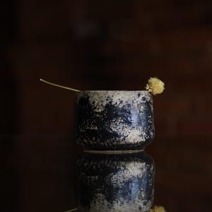 Puede incluir: Una peque&ntilde;a taza cil&iacute;ndrica de cer&aacute;mica con un acabado texturizado y moteado en negro y beige. Un tallo fino y claro con una peque&ntilde;a flor seca amarilla y redonda est&aacute; colocado apoyado contra el borde de la taza.