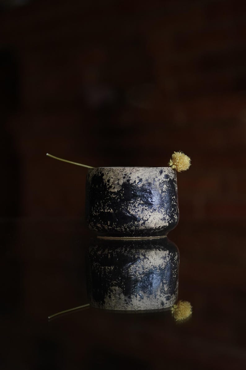 Puede incluir: Una peque&ntilde;a taza cil&iacute;ndrica de cer&aacute;mica con un acabado texturizado y moteado en negro y beige. Un tallo fino y claro con una peque&ntilde;a flor seca amarilla y redonda est&aacute; colocado apoyado contra el borde de la taza.