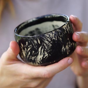 May include: A handcrafted ceramic bowl with a textured black exterior and a light interior. The bowl features a distinctive pattern of white brushstrokes, creating a striking contrast. The bowl is held in a person's hands.