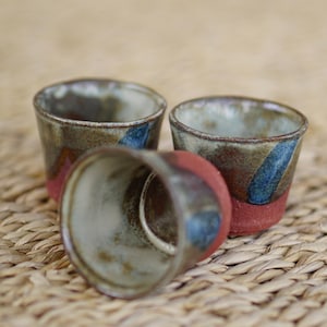 May include: Three small ceramic cups with a cylindrical shape. The exterior features a textured, reddish-brown base that transitions into a glazed upper section with mottled grey, white, and blue tones. The interior is a smooth, light cream colour.