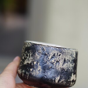 Puede incluir: Una peque&ntilde;a taza de cer&aacute;mica redonda que presenta un exterior negro mate con una textura pronunciada y parches desgastados de color arena que revelan la arcilla subyacente. El interior del recipiente es liso y de color blanco roto, contrastando con la superficie exterior oscura y rugosa.