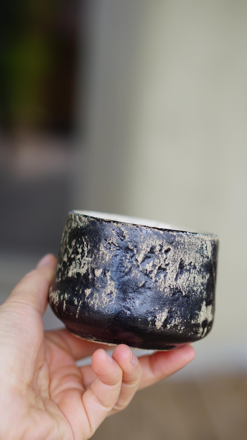 Puede incluir: Una peque&ntilde;a taza de cer&aacute;mica redonda que presenta un exterior negro mate con una textura pronunciada y parches desgastados de color arena que revelan la arcilla subyacente. El interior del recipiente es liso y de color blanco roto, contrastando con la superficie exterior oscura y rugosa.