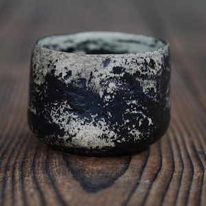 Puede incluir: Una peque&ntilde;a taza de cer&aacute;mica redonda con una superficie texturizada e irregular y un acabado de alto contraste. El exterior muestra un patr&oacute;n moteado de negro profundo y blanco roto, creando un aspecto rugoso con hendiduras visibles.