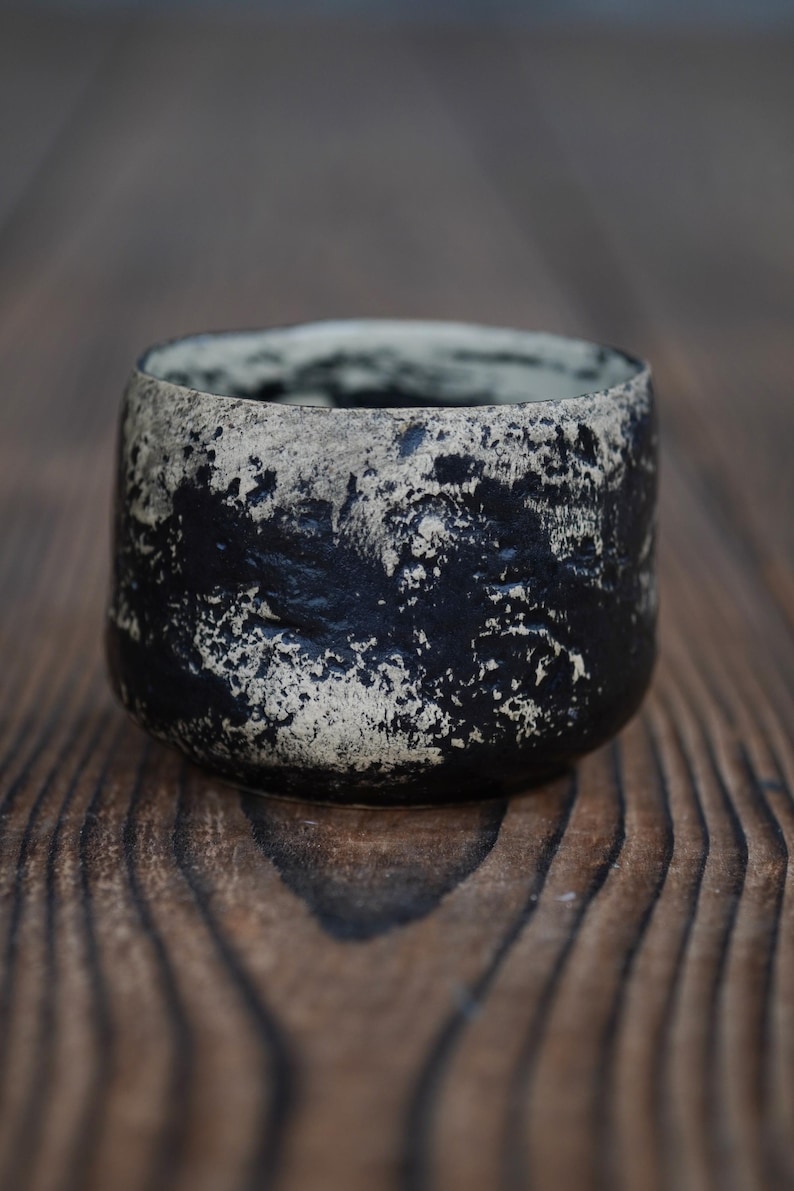 Puede incluir: Una peque&ntilde;a taza de cer&aacute;mica redonda con una superficie texturizada e irregular y un acabado de alto contraste. El exterior muestra un patr&oacute;n moteado de negro profundo y blanco roto, creando un aspecto rugoso con hendiduras visibles.