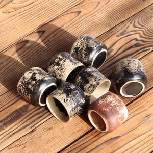 Puede incluir: Un conjunto de siete tazas peque&ntilde;as de cer&aacute;mica con forma cil&iacute;ndrica. Seis de las tazas presentan un exterior negro con un acabado beige texturizado y desgastado, mientras que una taza tiene un exterior marr&oacute;n rojizo con textura beige.