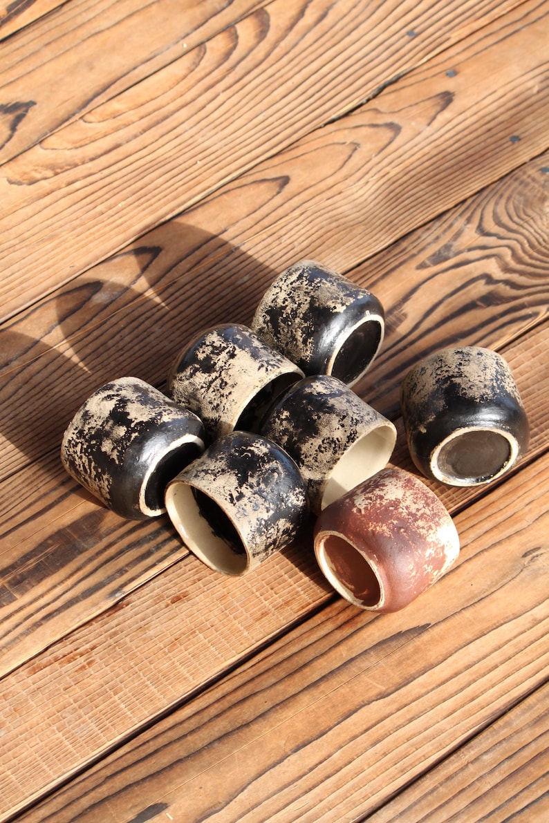 Puede incluir: Un conjunto de siete tazas peque&ntilde;as de cer&aacute;mica con forma cil&iacute;ndrica. Seis de las tazas presentan un exterior negro con un acabado beige texturizado y desgastado, mientras que una taza tiene un exterior marr&oacute;n rojizo con textura beige.