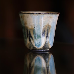May include: A handcrafted ceramic cup with a distinctive glaze. The cup has a light blue and tan colour scheme with dark blue accents. The cup's design is reflected in the glossy surface below. The cup is a unique piece of pottery.