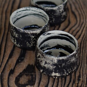 Puede incluir: Tres tazas peque&ntilde;as de cer&aacute;mica con un exterior negro texturizado y detalles en blanco moteado. El interior de cada taza es de color blanco roto y est&aacute; decorado con trazos de pincel negros que rodean las paredes interiores.