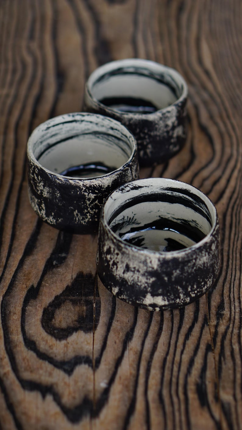 Puede incluir: Tres tazas peque&ntilde;as de cer&aacute;mica con un exterior negro texturizado y detalles en blanco moteado. El interior de cada taza es de color blanco roto y est&aacute; decorado con trazos de pincel negros que rodean las paredes interiores.