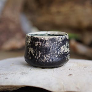 Puede incluir: Una peque&ntilde;a taza de cer&aacute;mica cil&iacute;ndrica con un exterior negro oscuro y un acabado blanco texturizado y moteado. El borde es liso y circular, mientras que el cuerpo tiene una base ligeramente c&oacute;nica con una apariencia rugosa e irregular.