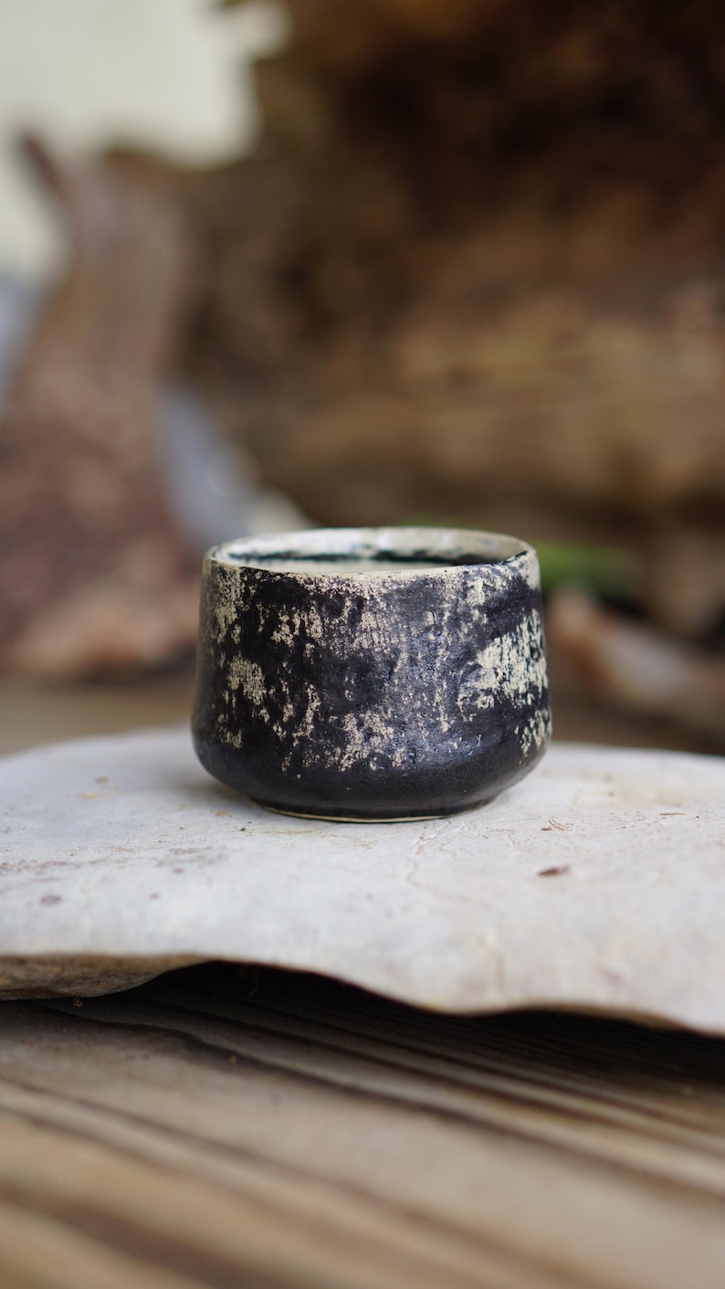 Puede incluir: Una peque&ntilde;a taza de cer&aacute;mica cil&iacute;ndrica con un exterior negro oscuro y un acabado blanco texturizado y moteado. El borde es liso y circular, mientras que el cuerpo tiene una base ligeramente c&oacute;nica con una apariencia rugosa e irregular.