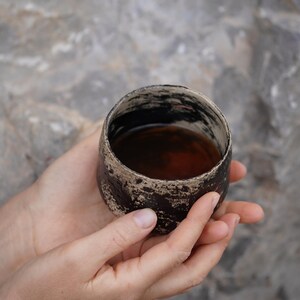 Puede incluir: Una peque&ntilde;a taza de cer&aacute;mica redonda con un acabado exterior texturizado en marr&oacute;n oscuro y negro. El borde de la taza es irregular y de color claro, contrastando con el cuerpo oscuro. El interior est&aacute; lleno de una bebida l&iacute;quida marr&oacute;n oscuro.