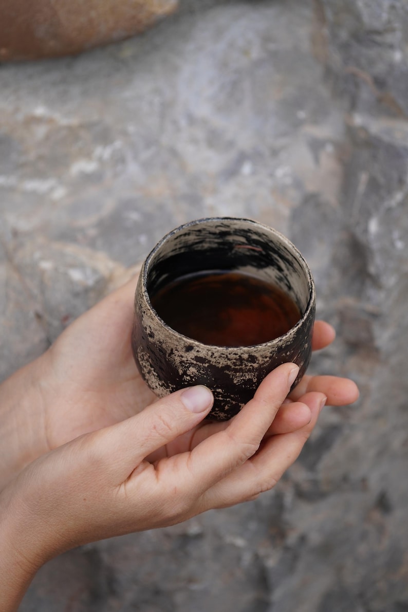 Puede incluir: Una peque&ntilde;a taza de cer&aacute;mica redonda con un acabado exterior texturizado en marr&oacute;n oscuro y negro. El borde de la taza es irregular y de color claro, contrastando con el cuerpo oscuro. El interior est&aacute; lleno de una bebida l&iacute;quida marr&oacute;n oscuro.