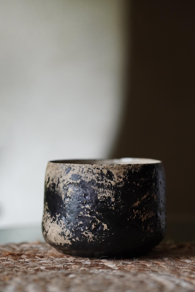 Puede incluir: Una peque&ntilde;a taza cil&iacute;ndrica de cer&aacute;mica con una superficie rugosa y muy texturizada. El exterior muestra un esquema de color contrastante de negro profundo y beige terroso, con un acabado moteado e irregular en toda la pared exterior.