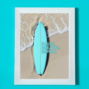 Könnte beinhalten: Ein gerahmter Druck mit einem hellblauen Surfbrett, das an einem Sandstrand mit sanften Wellen liegt. Das Kunstwerk enthält den Text "PAUSE. BREATHE. REPEAT." in Türkis und Blau. Der Rahmen ist weiß.