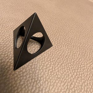 May include: A black, triangular metal object with two circular cutouts. The object is positioned on a textured, tan surface. The design is geometric and modern, suitable for home decor or display.