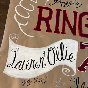 Op de afbeelding: Een bruin papieren bord met witte en kastanjebruine letters. Het bord zegt "Aggie Ring" en "Lauren & Ollie" in cursief. Een wit handgebaar en een ster zijn ook inbegrepen. De zin "gig 'em!" staat onderaan.