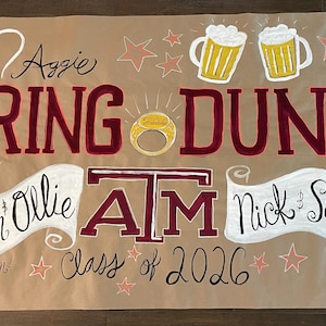 Puede incluir: Un letrero de papel marrón con las palabras "RING DUNK" en letras granates. El letrero incluye los nombres Lauren & Ollie y Nick & Sophie, con el texto "Class of 2026". Hay ilustraciones de jarras de cerveza, estrellas y un anillo.