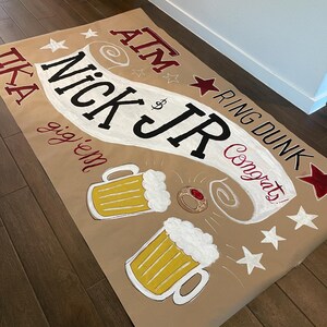Op de afbeelding: Een bruine papieren banner met de tekst "Nick Jr" in grote zwarte letters, omringd door sterren en bierpullen. De banner bevat ook de woorden "ATM", "Ring Dunk" en "Congrats!"