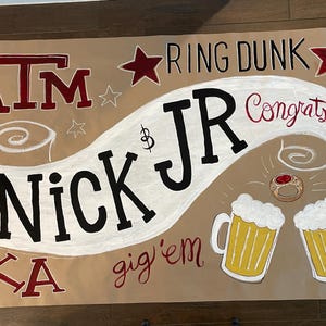 Op de afbeelding: Een bruine banner met kastanjebruine en zwarte letters. De banner zegt "ATM", "Nick Jr", "Ring Dunk", "Congrats!" en "gig'em". Er zijn ster- en bierkruikillustraties.