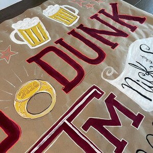 Op de afbeelding: Een bruine papieren banner met handgeschilderde elementen. Het bevat twee bierpullen, een gouden ring met "2026" erop en het woord "DUNK THEM" in bordeauxrood met witte contouren. Er zijn ook sterrenvormen op de achtergrond.