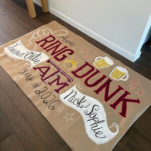 Op de afbeelding: Een grote, bruine banner met handgeschilderde tekst en illustraties. De banner bevat de woorden "RING DUNK" in bordeauxrood, met "Aggie" erboven. Andere tekst bevat namen en "Class of 2026". Bierpullen, sterren en een ring zijn ook afgebeeld.