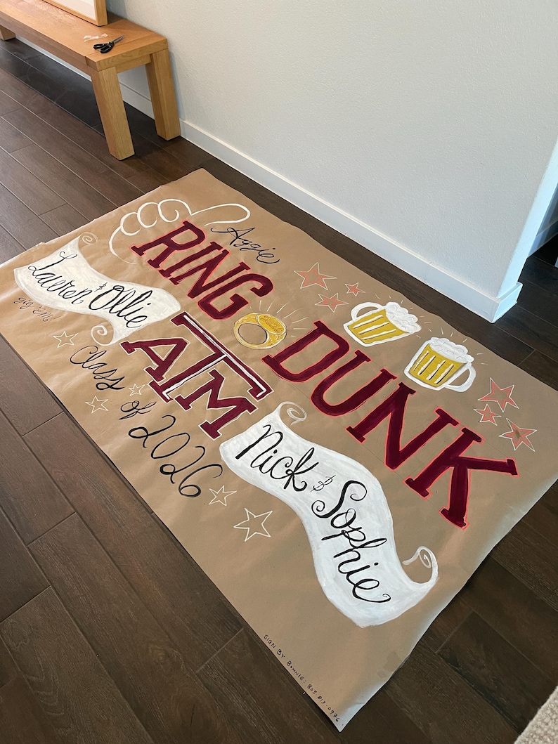Op de afbeelding: Een grote, bruine banner met handgeschilderde tekst en illustraties. De banner bevat de woorden "RING DUNK" in bordeauxrood, met "Aggie" erboven. Andere tekst bevat namen en "Class of 2026". Bierpullen, sterren en een ring zijn ook afgebeeld.