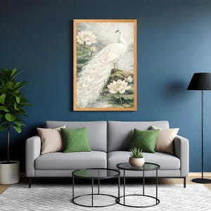 May include: A framed watercolour painting of a white peacock with a long, flowing tail, standing on a rock near water lilies. The artwork has a light wooden frame and is displayed on a blue wall above a grey sofa with green and beige cushions.