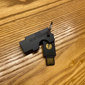 Może przedstawiać: Czarny pendrive z kółkiem na klucze, z napisem "MAX MUSTERMANN" i złotym okręgiem z symbolem "Y". Pendrive leży na drewnianej powierzchni.