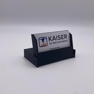 Könnte beinhalten: Ein schwarzer Visitenkartenhalter mit rechteckiger Basis und abgewinkeltem Kartenanzeigebereich. Der Halter zeigt eine Visitenkarte mit dem Text "KAISER 3D DRUCKFABRIK" und einer 3D-Drucker-Grafik. Der Hintergrund ist weiß.