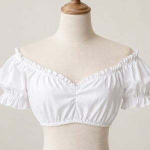 Op de afbeelding: Een witte off-the-shoulder crop top met korte, gepofte mouwen. De top heeft een gerimpelde halslijn en mouwboorden, en een geplooid detail bij de buste. De stof lijkt een lichtgewicht katoen of iets dergelijks te zijn.