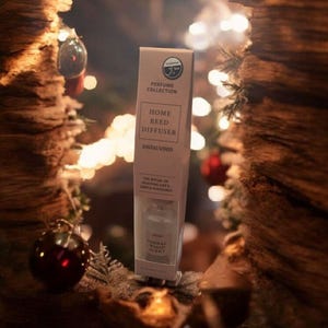 May include: A beige Home Reed Diffuser box from the Perfume Collection, with the text "Sandalwood". The box contains a glass bottle with reeds. The background features a Christmas tree with baubles and fairy lights.