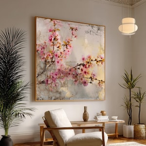 Può includere: Grande opera d'arte incorniciata con rami di fiori di ciliegio rosa. Il dipinto ha uno sfondo strutturato e astratto con toni neutri e una cornice dorata. L'opera d'arte è esposta in una stanza ben illuminata con piante e mobili.