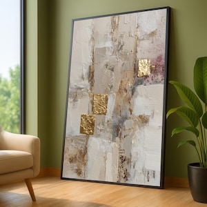 May include: Large abstract painting in a black frame. The artwork features a textured surface with a palette of beige, white, and brown tones, accented by gold leaf squares. The painting leans against a green wall.