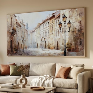 Puede incluir: Una pintura al óleo representa una escena de calle europea con edificios en tonos beige y marrón. La obra de arte presenta una calle con edificios, farolas y figuras. La pintura está enmarcada en un marco de madera clara y cuelga sobre un sofá.