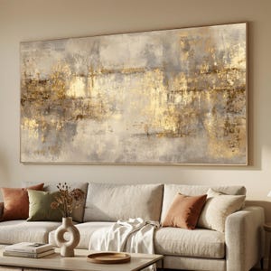 Puede incluir: Gran pintura abstracta con una paleta de colores dorados y grises. La obra de arte presenta pinceladas texturizadas y un marco rectangular dorado. La pintura cuelga sobre un sofá beige con cojines decorativos.