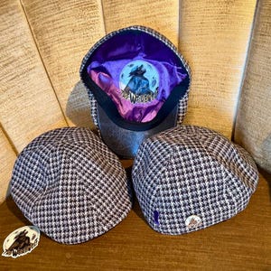 Op de afbeelding: Drie petten met een pied-de-poule patroon. Eén pet is open en onthult een paarse binnenkant met een weerwolfafbeelding en de tekst "The Werewolf". Er is ook een sticker met dezelfde afbeelding.