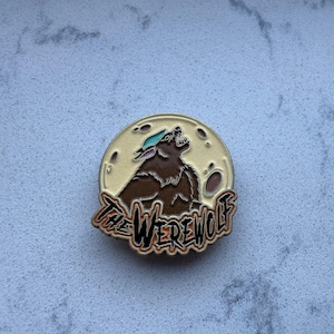 May include: A round pin featuring a brown werewolf howling at a full moon. The pin has a cream-colored background with the words "The Werewolf" in a stylized font. The werewolf has a teal accent on its head.