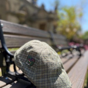 May include: A green and brown plaid newsboy cap with a small embroidered patch that reads "CREATURE". The cap is resting on a wooden bench. The cap has a classic, flat design.