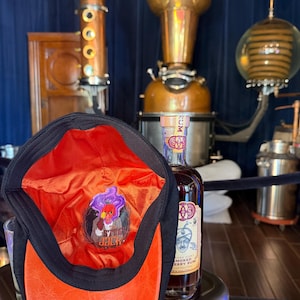 May include: A black cap with a bright orange interior lining featuring a graphic print of a pumpkin-headed character with purple smoke and the text Stingy Jack. The cap is positioned next to a glass bottle of rum with a label featuring a man's face.