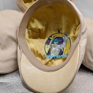 May include: A beige flat cap with a textured brim and a gold-colored satin lining. The interior features a circular graphic print of a mummy character with the text THE MUMMY in blue letters. The cap has a structured crown and a stitched panel design.