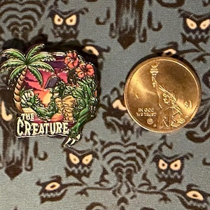 May include: Enamel pin featuring "The Creature" from a tropical scene with palm trees and flowers. The pin is next to a US quarter for size comparison. The pin has green, purple, and orange colors.